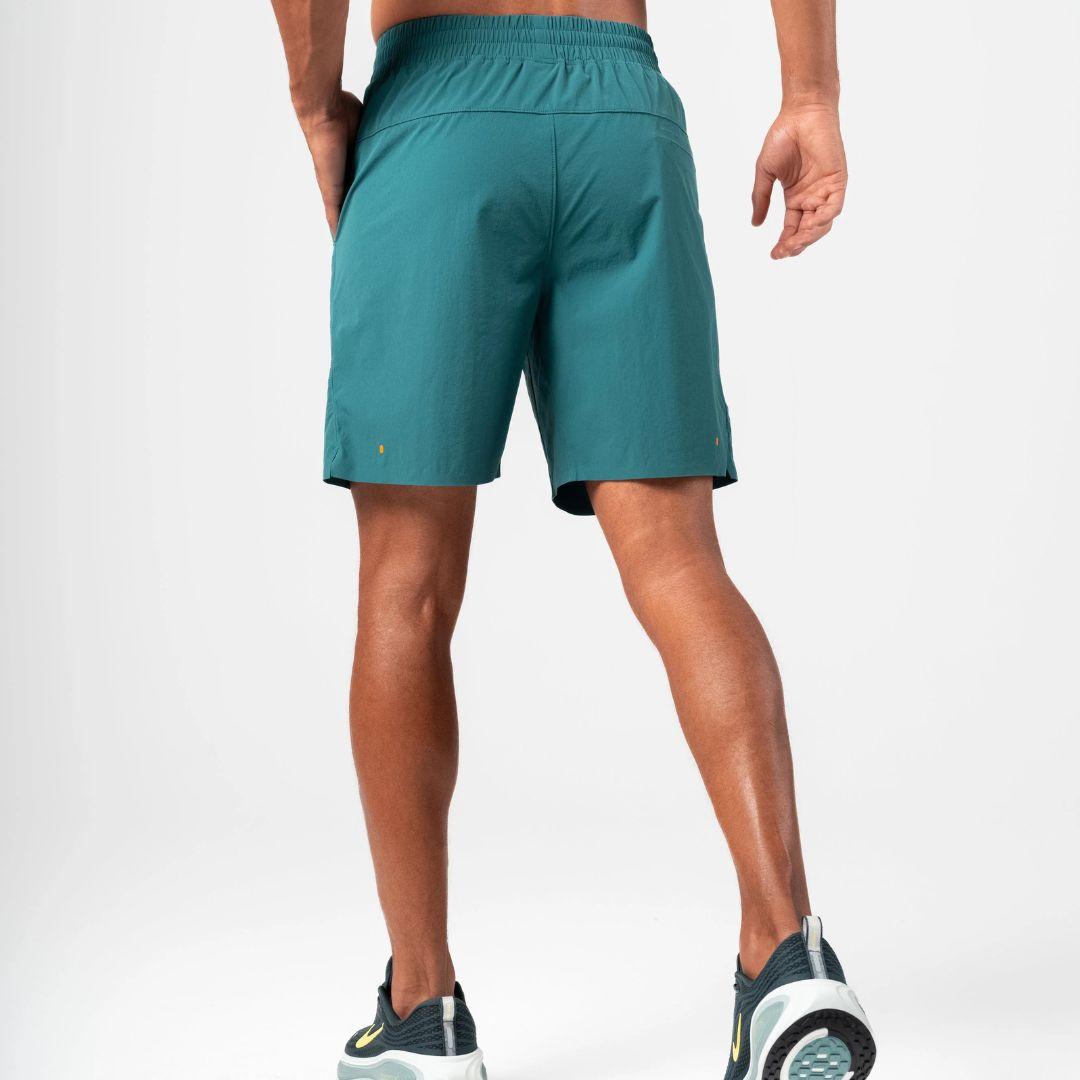 A man wearing teal athletic shorts and dark running shoes, shown from the back against a clean white studio background.