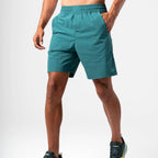 A man wearing teal athletic Agaru sports shorts and dark running shoes, shown from the waist down against a light gray studio background.