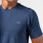 Close-up of a man wearing a heathered blue short-sleeve athletic t-shirt with a light blue geometric Agaru Sports logo on the left chest, set against a plain light gray background.