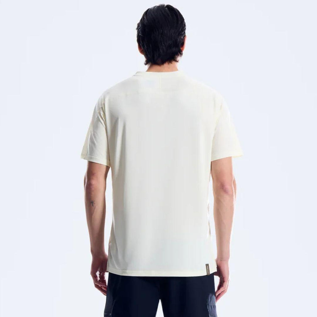 A man viewed from the back wearing a short-sleeve crewneck t-shirt in a warm off-white/cream color, paired with dark pants, against a clean white studio background.