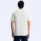 A man viewed from the back wearing a short-sleeve crewneck t-shirt in a warm off-white/cream color, paired with dark pants, against a clean white studio background.