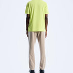 A man viewed from the back wearing a bright lime green t-shirt and beige trousers, standing against a plain white studio background.