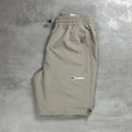 A pair of light Beige Berghaus shorts laid flat on a textured gray concrete surface. The shorts feature an elastic waistband with a black drawstring, a zippered pocket on the right side, and the Berghaus logo embroidered in white, red, and blue on the left thigh.