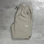 A pair of light Beige Berghaus shorts laid flat on a textured gray concrete surface. The shorts feature an elastic waistband with a black drawstring, a zippered pocket on the right side, and the Berghaus logo embroidered in white, red, and blue on the left thigh.