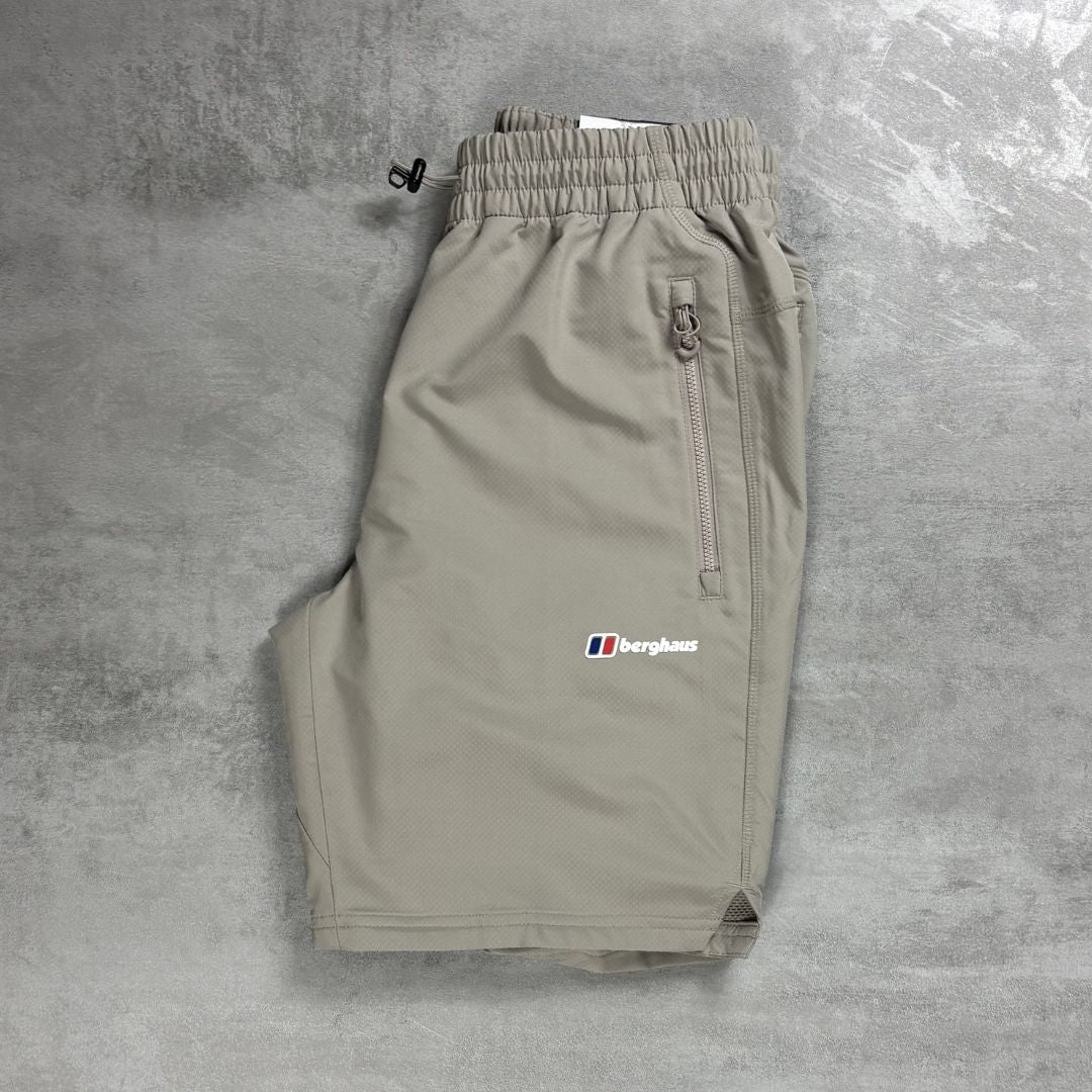 A pair of light Beige Berghaus shorts laid flat on a textured gray concrete surface. The shorts feature an elastic waistband with a black drawstring, a zippered pocket on the right side, and the Berghaus logo embroidered in white, red, and blue on the left thigh.