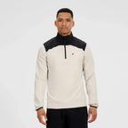 A man wearing a Berghaus long-sleeve athletic pullover in off-white and black, featuring a black mock neck and shoulder yoke, a black half-zip collar, and a small red and blue logo on the left chest. He is standing against a solid light gray background.