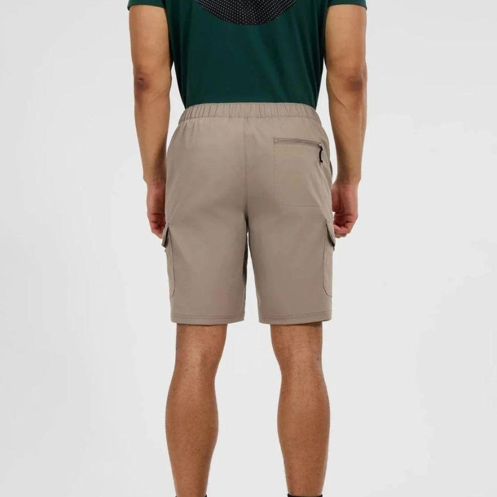 A man viewed from the back wearing beige cargo shorts and a dark green t-shirt, standing against a light gray studio background.