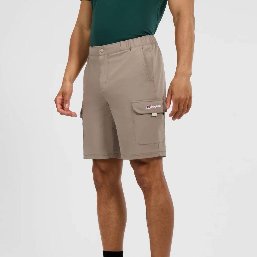 A man wearing beige cargo shorts with a green t-shirt, shown from the waist down against a light gray studio background. The shorts feature a button closure, elastic waistband, and a cargo pocket on the right leg with a visible Berghaus logo.