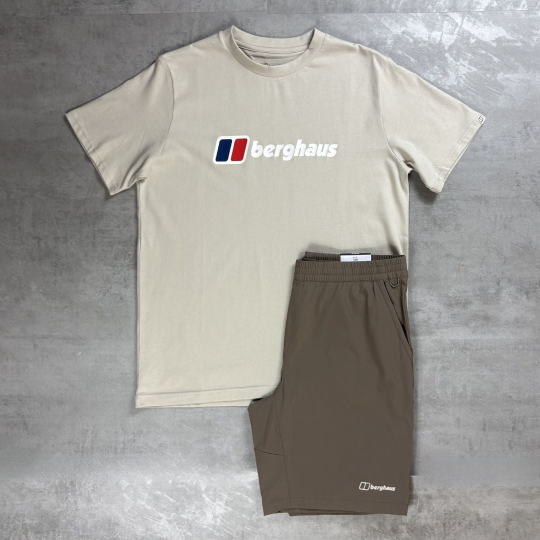 A beige short-sleeve t-shirt with a Berghaus logo on the chest, laid flat next to a pair of brown shorts on a gray concrete-textured background.