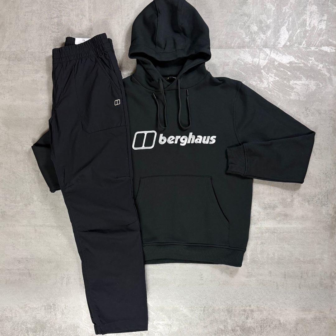 A black Berghaus hoodie and matching black jogger pants laid flat on a textured gray concrete surface. The hoodie features a white printed logo on the chest, and the joggers have an elastic waist and a small white logo on the left pocket.
