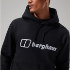 A man wearing a black pullover hoodie with a large silver and white Berghaus logo printed across the chest, shown against a neutral gray background.