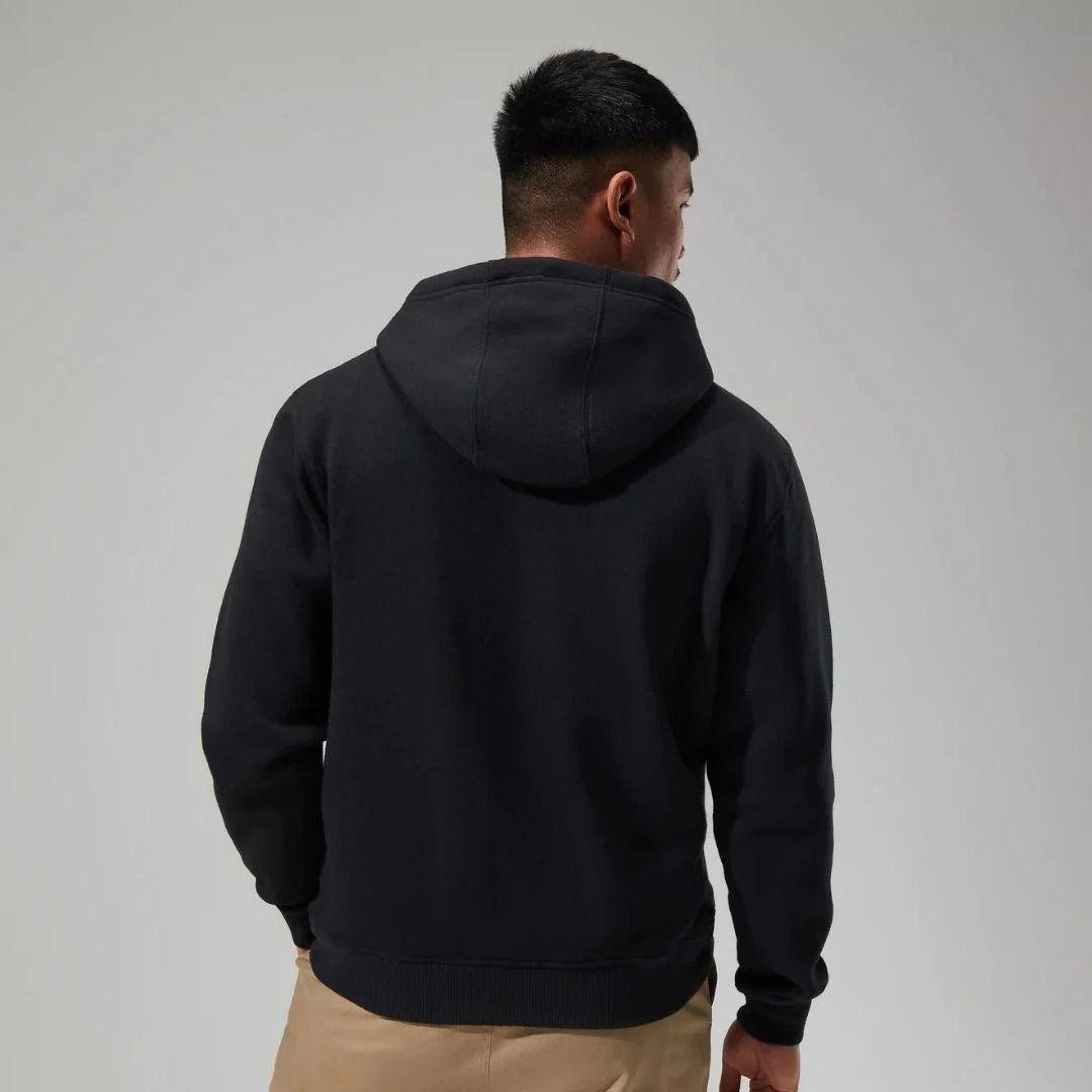 A man viewed from behind wearing a black pullover hoodie and tan pants against a plain light gray studio background.