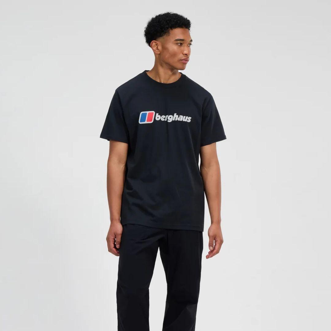 A man wearing a black Berghaus t-shirt with the brand's logo printed across the chest, paired with black trousers, photographed against a light gray studio background.