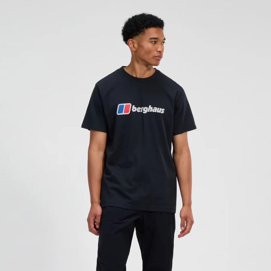 A man wearing a black short-sleeve crewneck t-shirt with a white and red 'berghaus' logo printed across the chest, standing against a light gray studio background.