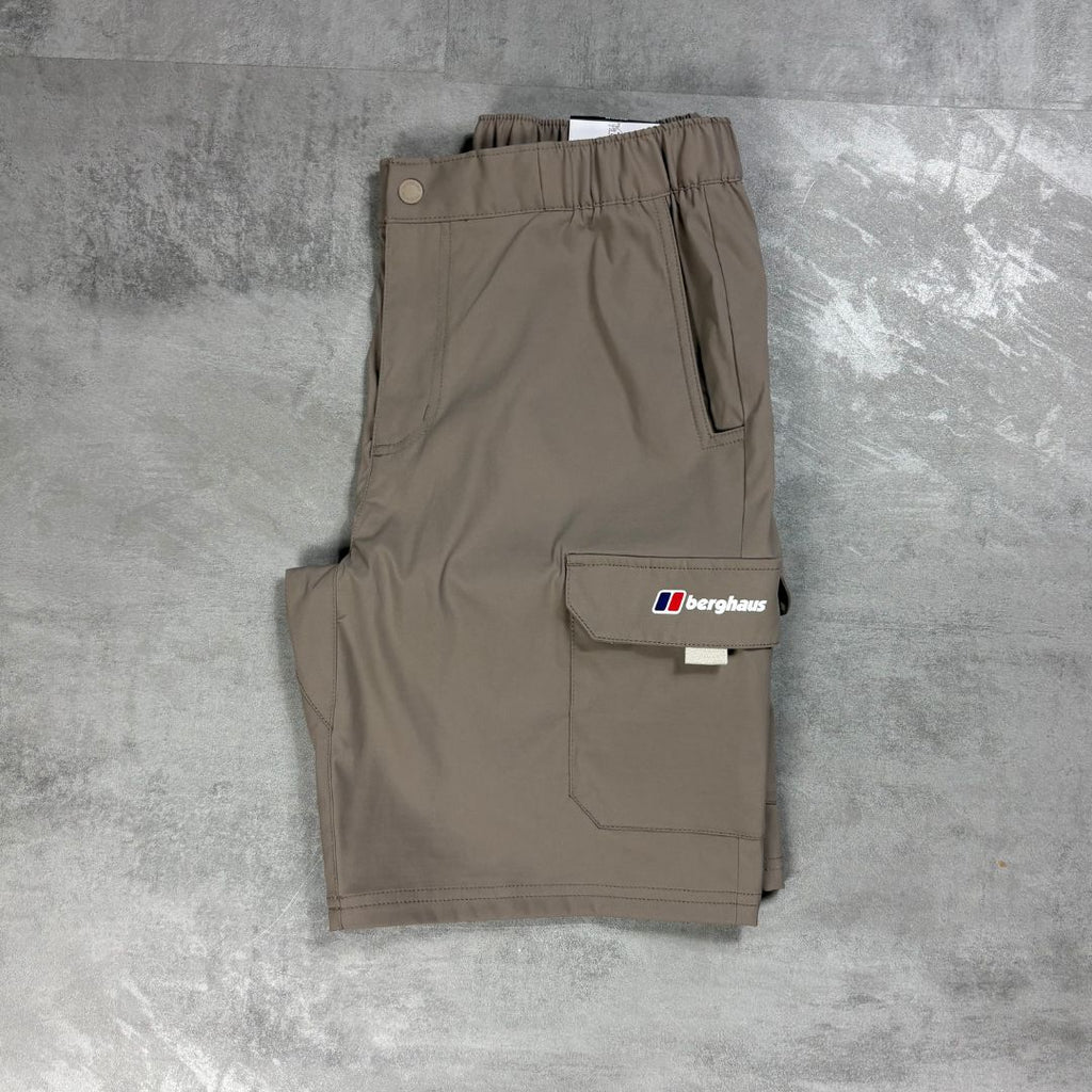 A pair of folded beige cargo shorts with an elastic waistband, laid flat on a textured gray concrete surface. The shorts feature a cargo pocket on the left leg with a white and red Berghaus logo patch.