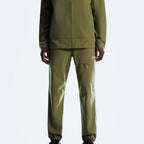 A person wearing an Berghaus olive green zip-front jacket and matching jogger-style pants with a small multicolor Berghaus logo patch on the upper thigh, photographed against a clean light gray studio background.