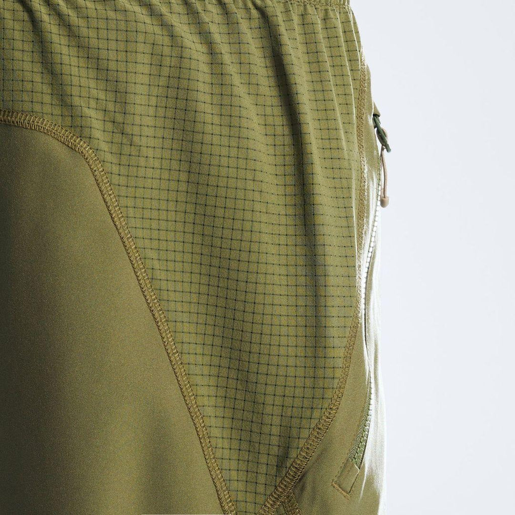 Close-up detail of olive green athletic shorts showing a fine grid patterned fabric panel, solid fabric side panel, and a zipper pocket on a white background.
