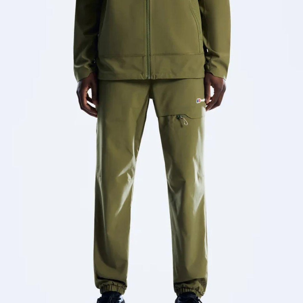 A person wearing olive green outdoor apparel, including a zip-front jacket and matching pants with a small multicolor logo patch on the upper thigh, photographed against a light gray studio background.