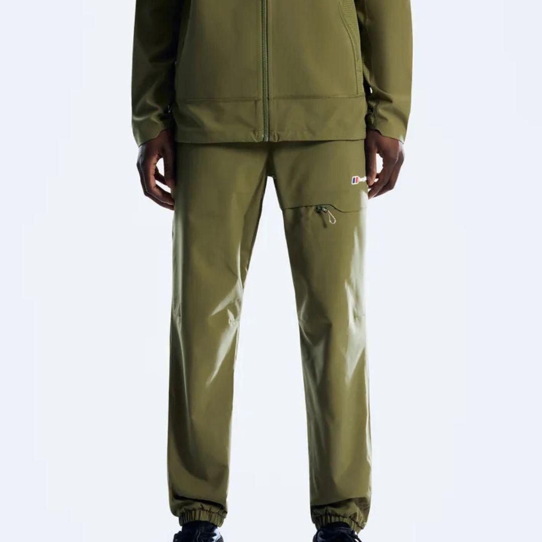 A person wearing olive green outdoor apparel, including a zip-front jacket and matching pants with a small multicolor logo patch on the upper thigh, photographed against a light gray studio background.