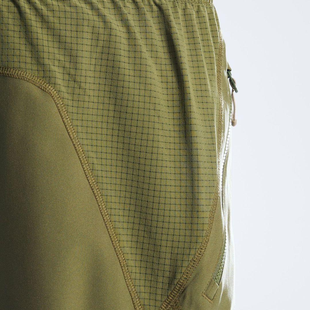 Close-up of olive green athletic shorts showing a fine grid pattern on the upper section, solid fabric on the lower section, and a side zipper.