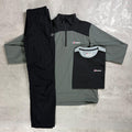A flat lay of a three-piece Berghaus outdoor clothing set on a textured gray concrete background. The set includes a black and gray quarter-zip pullover, matching black trousers, and a black short-sleeve T-shirt with a small Berghaus logo on the chest.