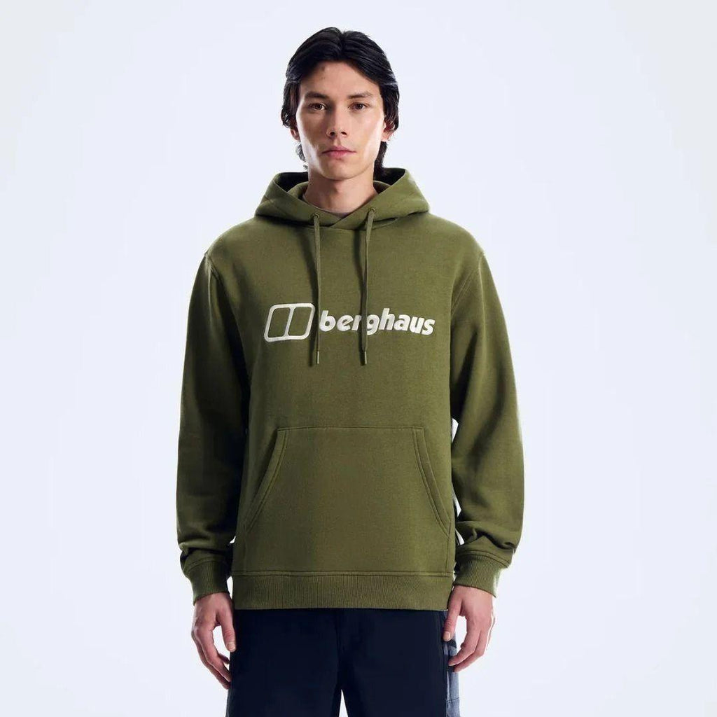 A man wearing an olive green pullover hoodie with a white printed 'berghaus' logo on the chest, paired with dark pants, photographed against a clean white studio background.