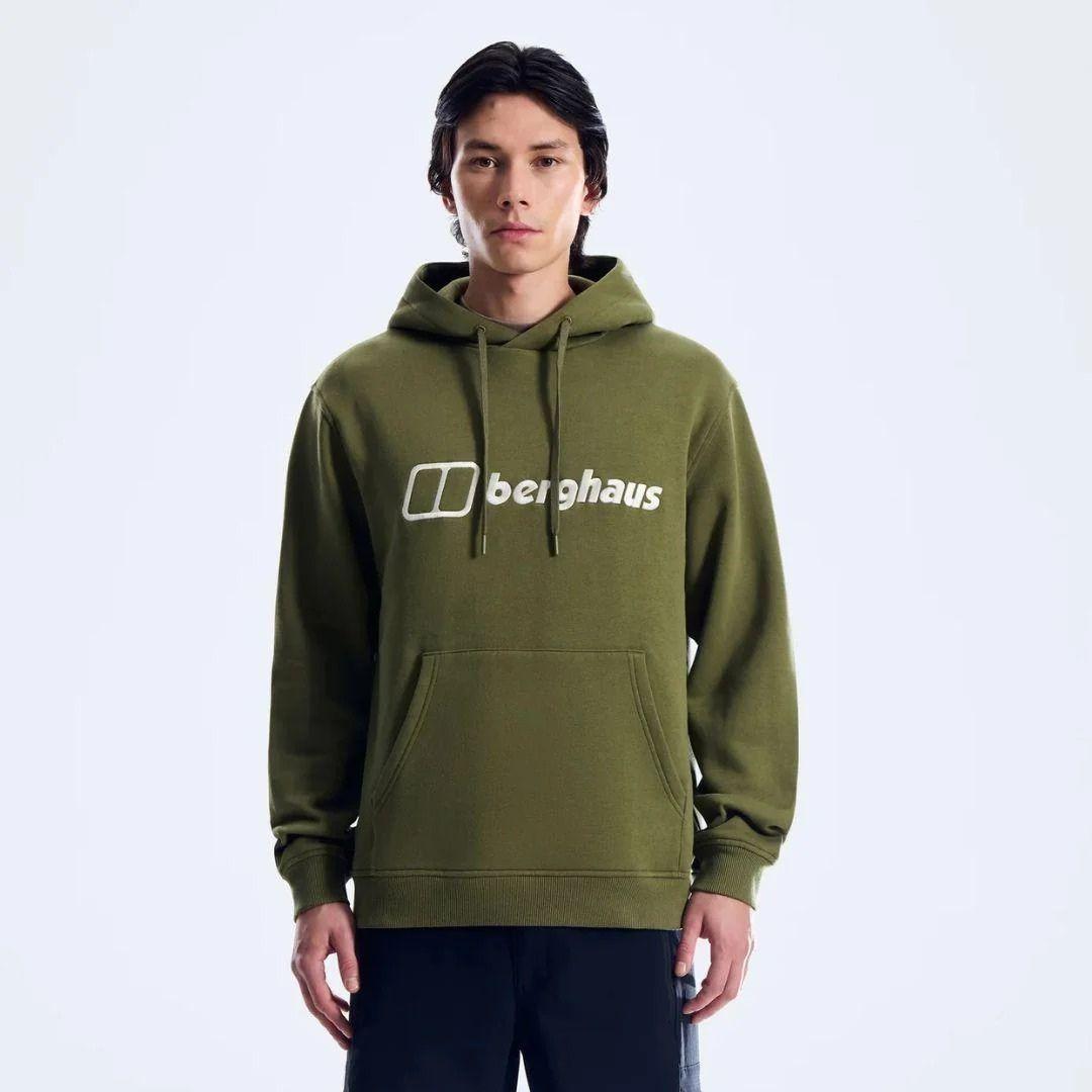A man wearing an olive green pullover hoodie with a white printed 'berghaus' logo on the chest, paired with dark pants, photographed against a clean white studio background.