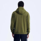 A man viewed from the back wearing an olive green pullover hoodie and dark pants against a plain light gray studio background.
