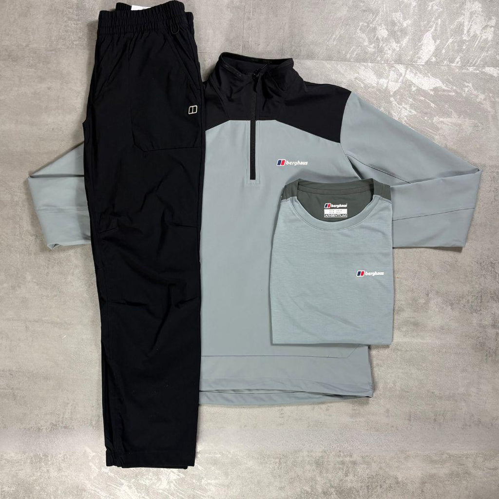 A three-piece men's athletic outfit laid flat on a gray textured surface, including black jogger pants, a light gray and black quarter-zip pullover, and a matching light gray t-shirt. The garments are branded with the Berghaus logo.