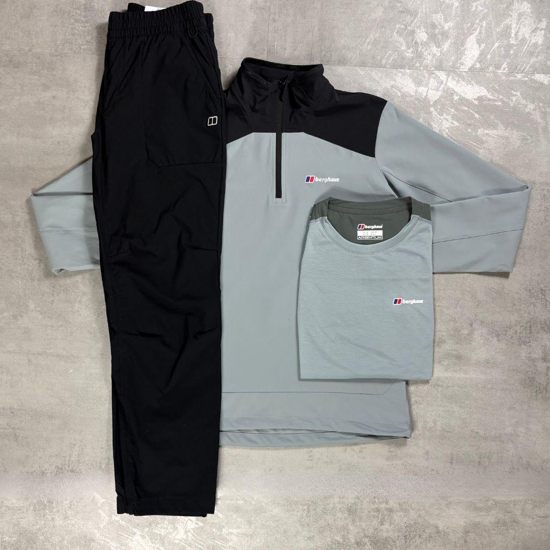 A three-piece men's athletic outfit laid flat on a gray textured surface, including black jogger pants, a light gray and black quarter-zip pullover, and a matching light gray t-shirt. The garments are branded with the Berghaus logo.