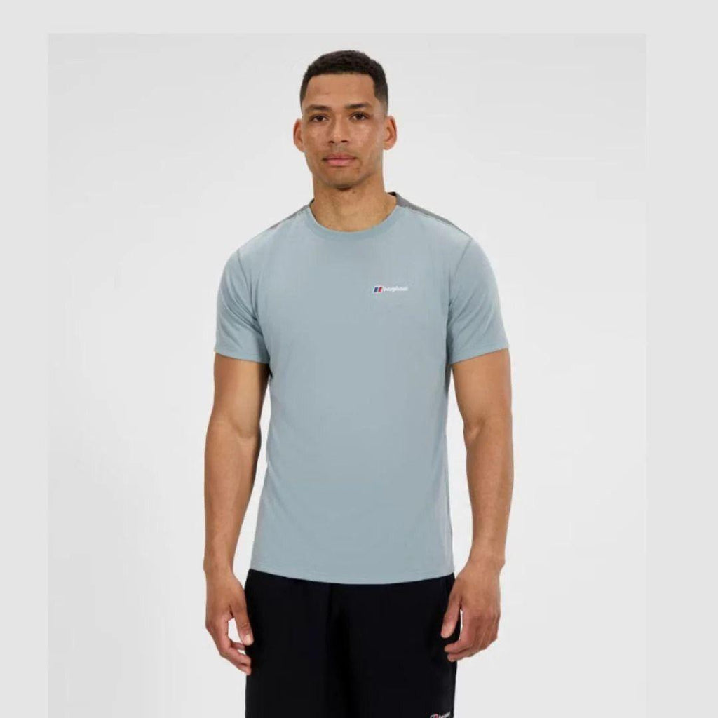 A man wearing a light blue athletic t-shirt with a Berghaus small red and white logo on the chest, paired with black shorts, standing against a light gray background.