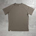 A short-sleeve Berghaus athletic t-shirt in a muted taupe-brown color, laid flat on a textured gray concrete surface. The shirt features raglan sleeves and a crew neck, with a small white square logo on the right sleeve cuff and a small white tag near the bottom hem.