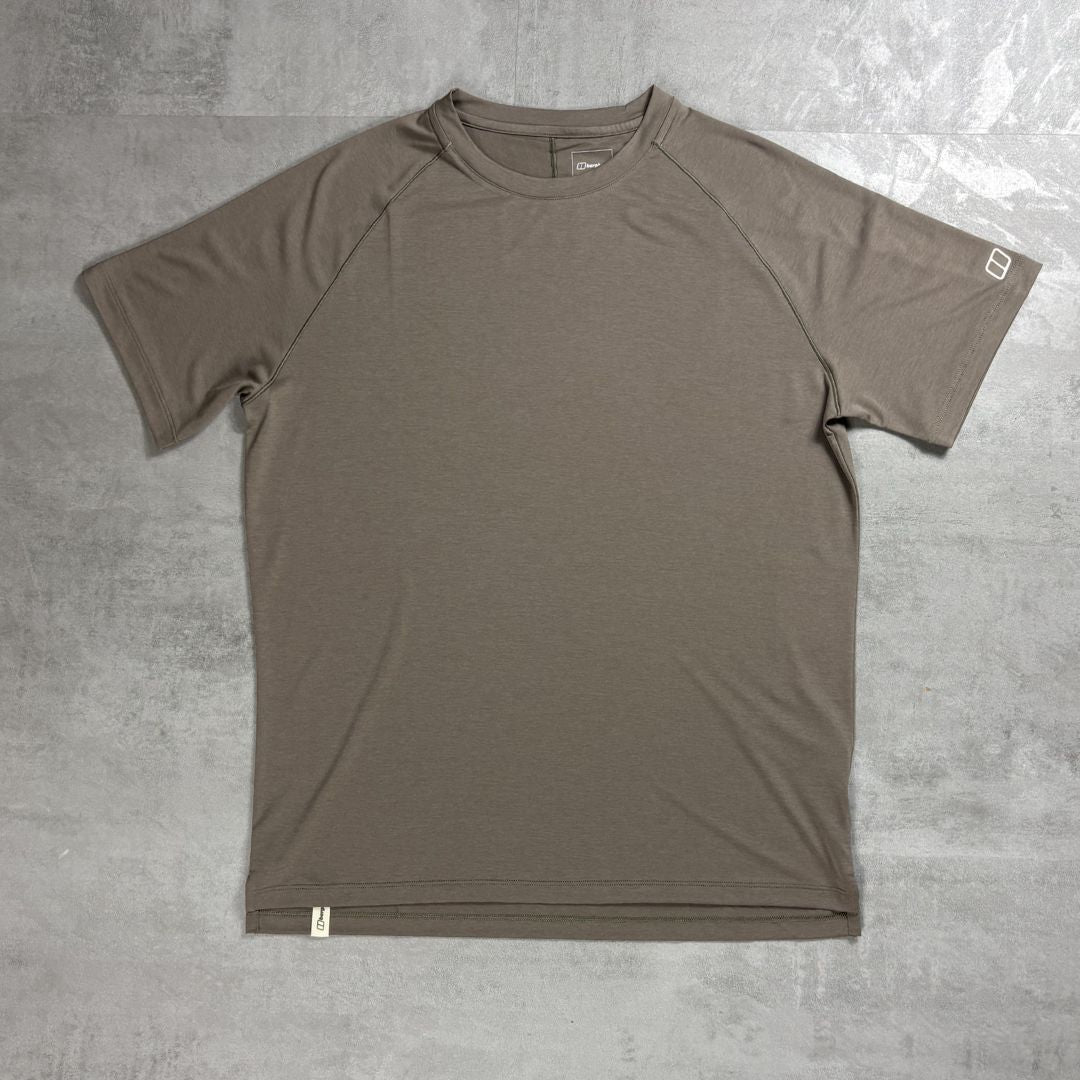 A short-sleeve Berghaus athletic t-shirt in a muted taupe-brown color, laid flat on a textured gray concrete surface. The shirt features raglan sleeves and a crew neck, with a small white square logo on the right sleeve cuff and a small white tag near the bottom hem.