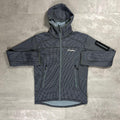 A dark gray hooded technical jacket with a subtle grid pattern, laid flat on a textured gray surface. The jacket features a full-length front zipper, two zippered hand pockets, a reflective arm patch on the upper sleeve, and a white Berghaus logo on the left chest.