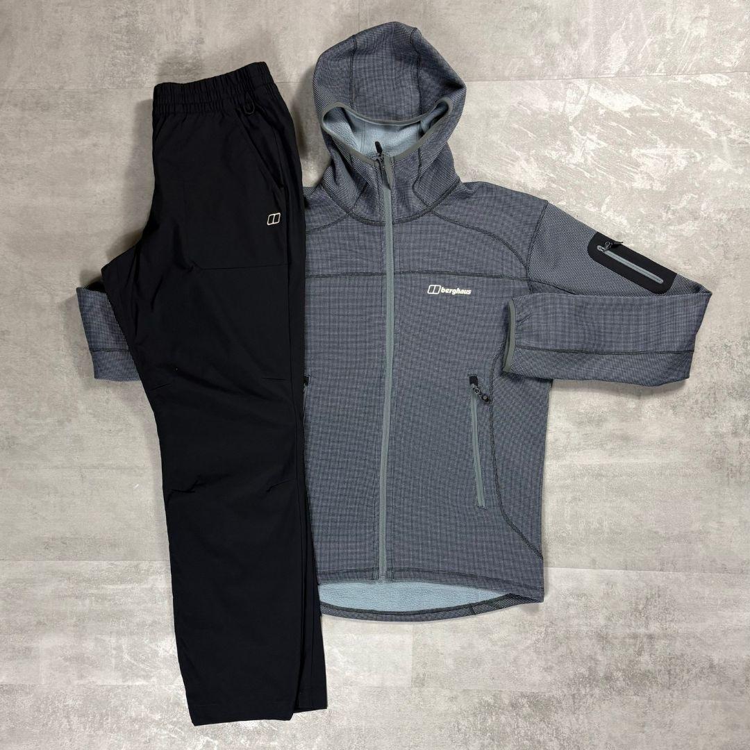 A flat-lay view of a two-piece outdoor apparel set consisting of a gray and black patterned hooded jacket and black pants, laid out on a gray textured floor. The brand name Berghaus is visible on the chest of the jacket.