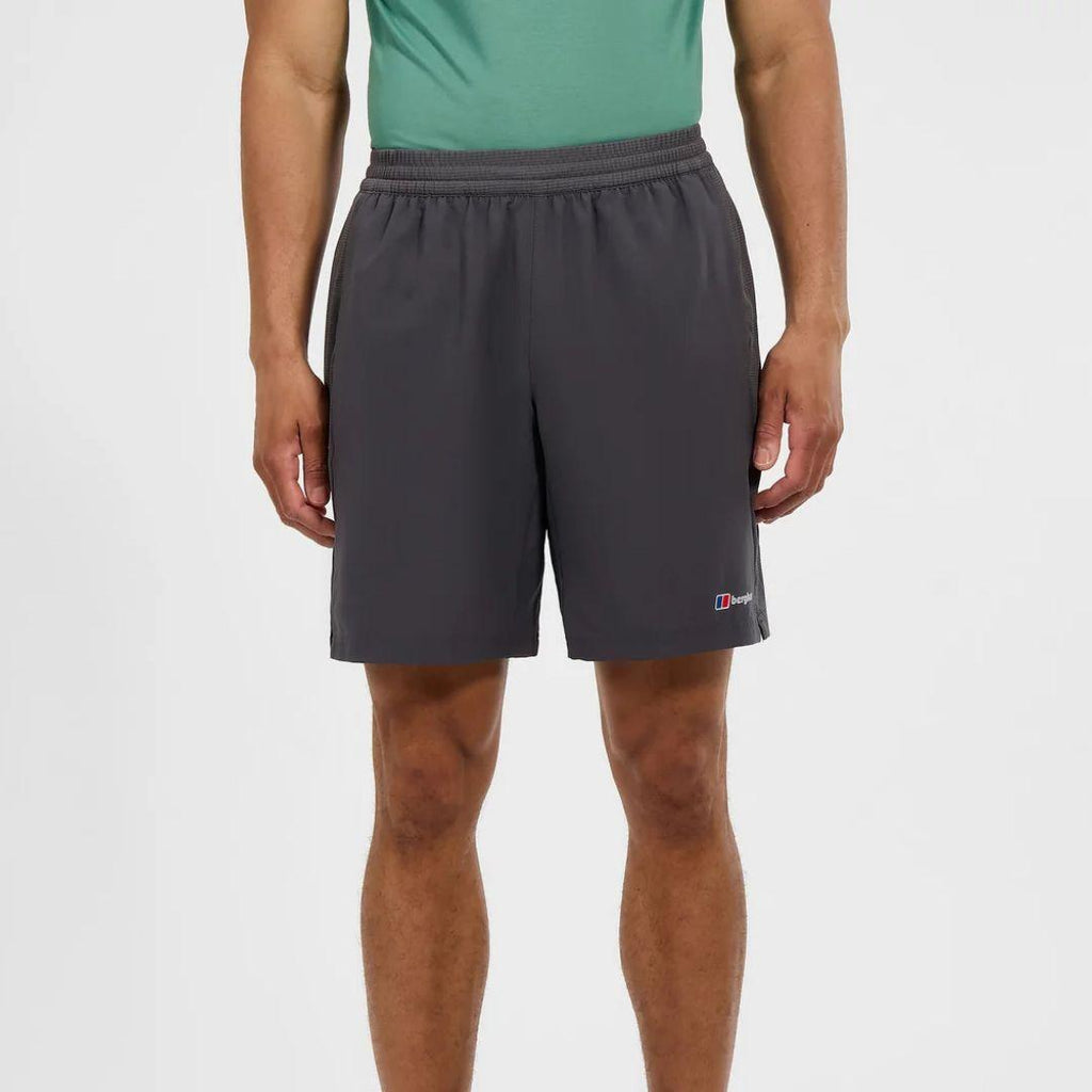 A man wearing dark gray athletic shorts and a green tank top, shown from the waist down against a plain light gray studio background. A small Berghaus logo is visible on the lower left leg of the shorts.