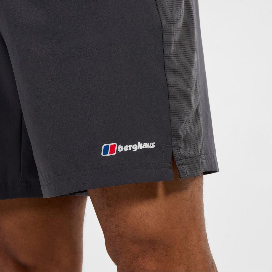 Close-up of a person wearing dark charcoal gray Berghaus athletic shorts, showing the side logo and the model's legs against a light gray background.