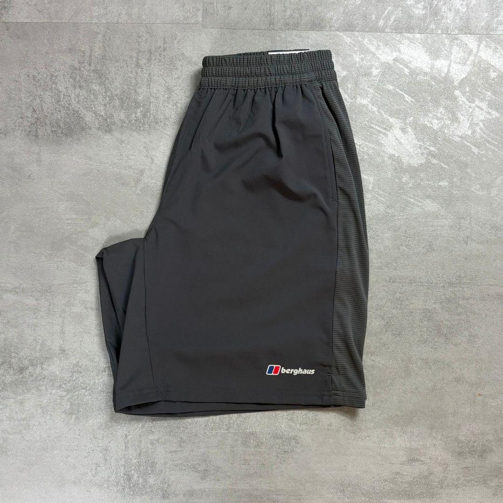 A pair of dark gray athletic shorts with an elastic waistband and a small Berghaus logo on the lower left leg, laid flat on a textured gray concrete surface.