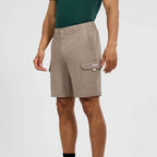 A man wearing beige cargo shorts with a green t-shirt, shown from the waist down against a light gray studio background. The shorts feature a button closure, elastic waistband, and a cargo pocket on the right leg with a visible Berghaus logo.