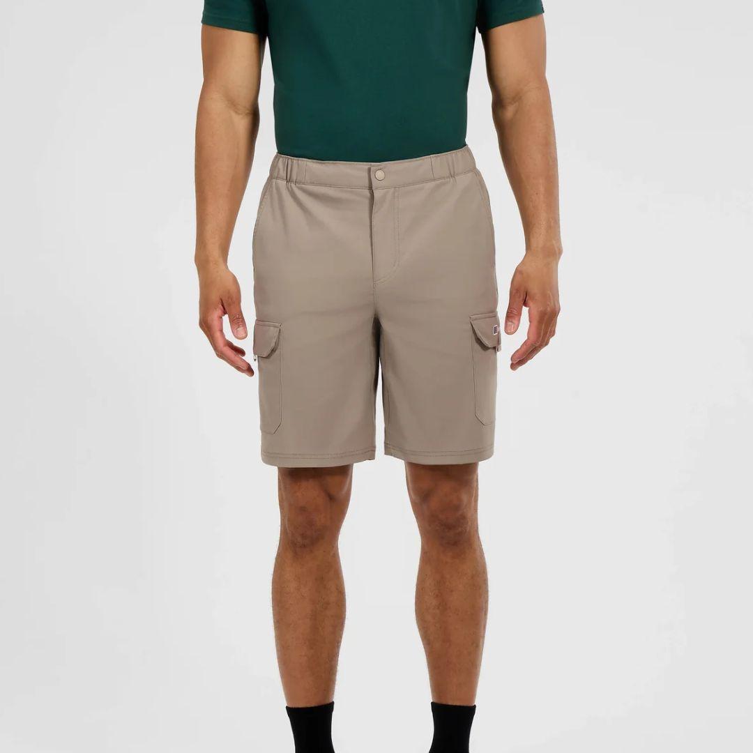 A man wearing beige cargo shorts with side flap pockets, paired with a dark green polo shirt and black socks, shown from the chest down against a light gray studio background.