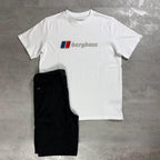 A white Berghaus short-sleeve t-shirt with the brand's logo printed on the chest, laid flat on a gray concrete surface next to a pair of black cargo pants.