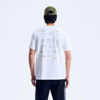 A man viewed from behind wearing a white t-shirt with a geometric line-art graphic print on the back, paired with black pants and an olive green baseball cap against a light gray studio background.
