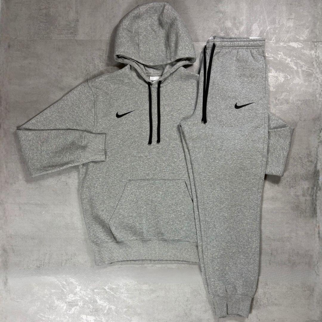 Nike Grey Club Fleece Hoodie and Joggers Tracksuit for men, flat lay of marl grey cotton fleece pullover hoodie with drawcord hood and kangaroo pocket and matching cuffed joggers with black Nike swoosh