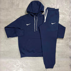Nike Navy Club Fleece Hoodie and Joggers Tracksuit for men, flat lay of navy cotton fleece pullover hoodie with white drawcords and kangaroo pocket and matching cuffed joggers with white Nike swoosh