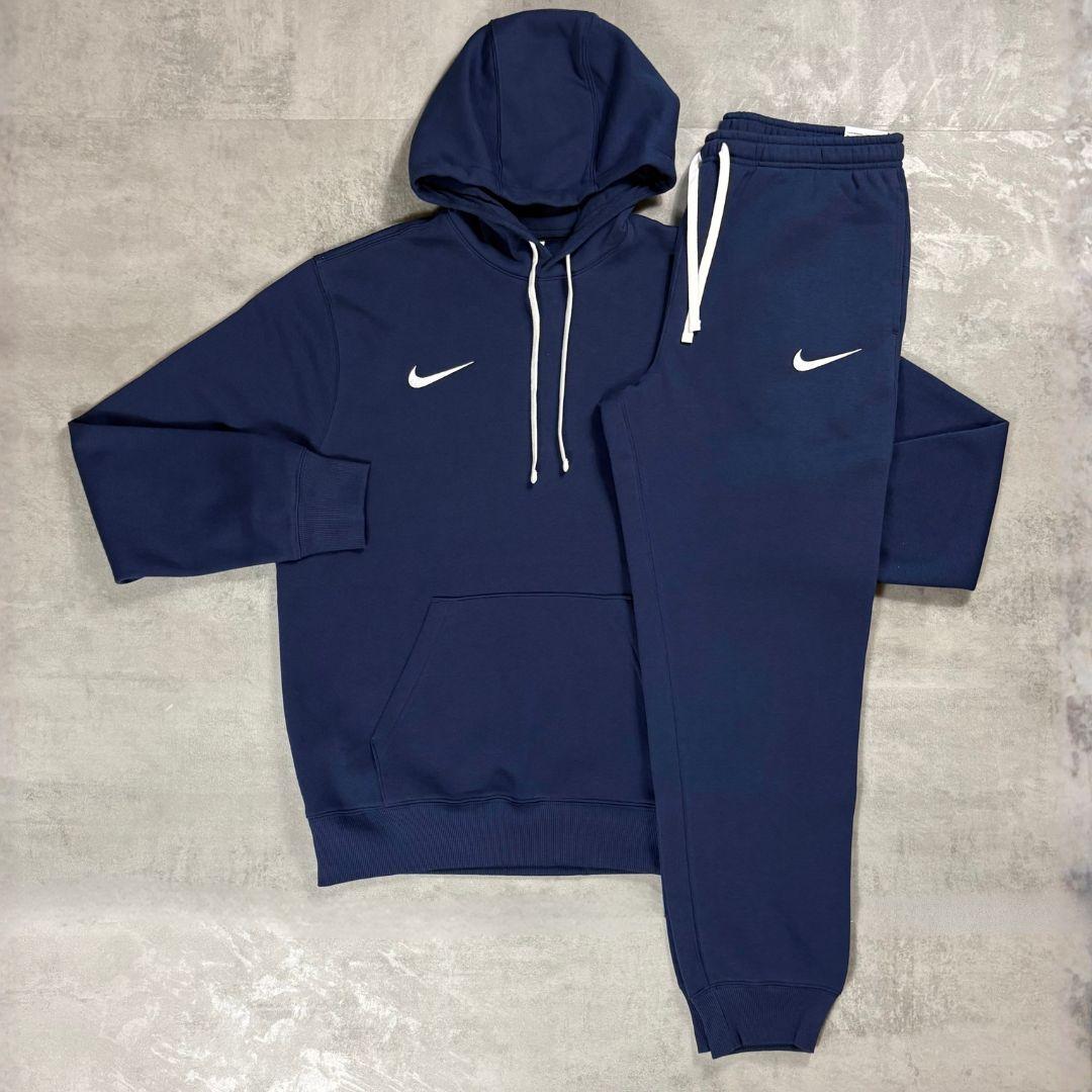 Nike Navy Club Fleece Hoodie and Joggers Tracksuit for men, flat lay of navy cotton fleece pullover hoodie with white drawcords and kangaroo pocket and matching cuffed joggers with white Nike swoosh
