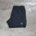 Grey athletic shorts with an elastic waistband and a small white Asics logo on the lower left leg, laid flat on a gray textured surface.